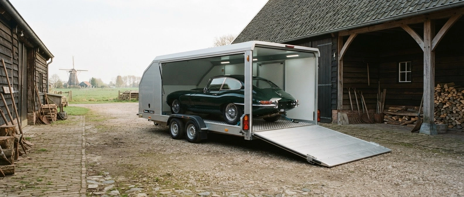 Groene klassieke sportwagen wordt in een gesloten witte Brian James-trailer geladen bij een boerenschuur